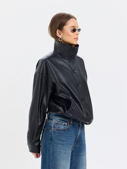 A woman wearing a black faux-leather jacket with a high collar and front buttons, paired with blue jeans, photographed against a plain light gray studio background.
