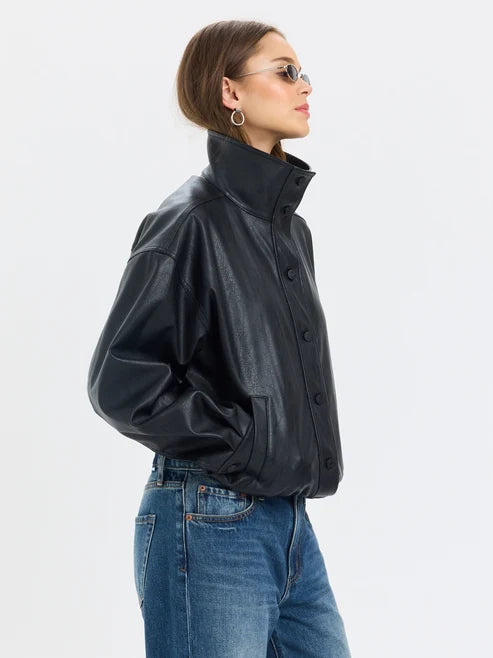 A woman in side profile wearing a black faux-leather jacket with a high collar and button front, paired with blue jeans, against a light gray studio background.
