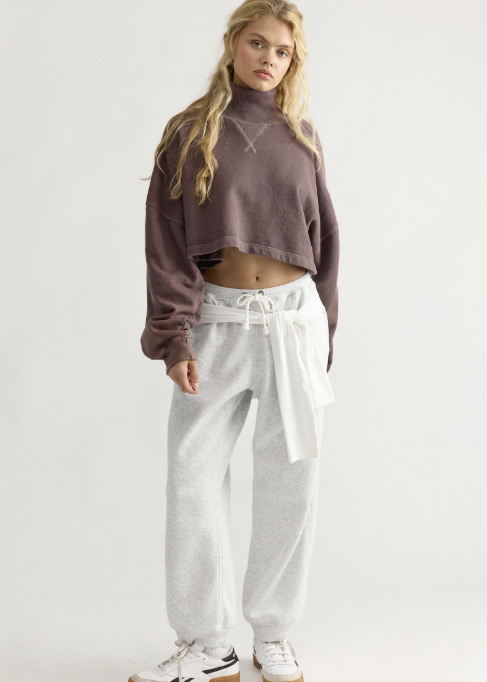 Woman wearing brown mock neck sweatshirt and grey sweatpants on white background