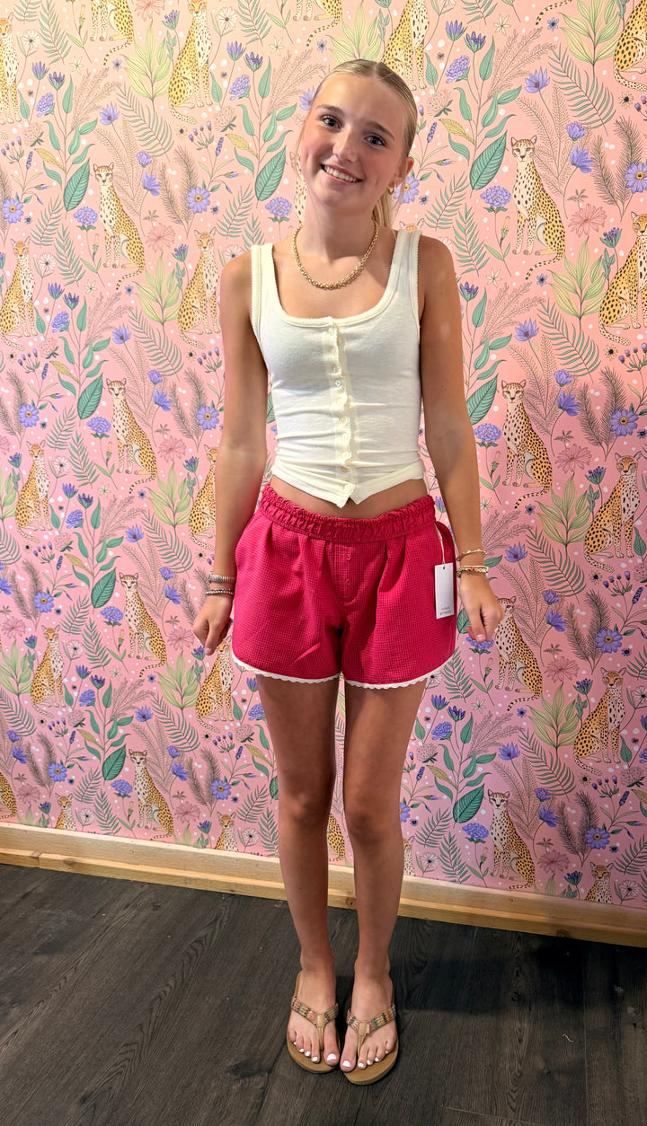 A young woman stands in front of a pink leopard-print wallpaper wall, wearing a white ribbed tank top with a button placket and bright pink shorts with white fringe.