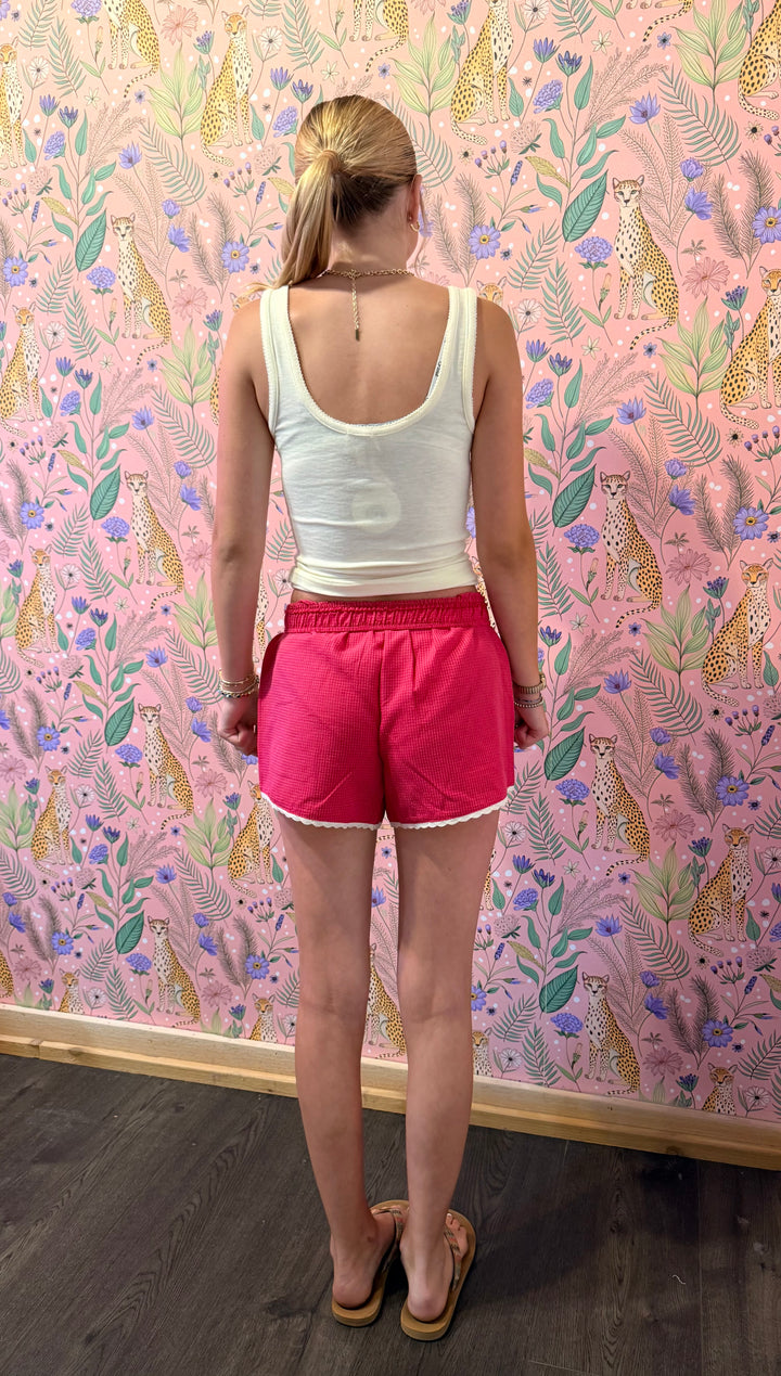 A woman stands with her back to the camera wearing a white tank top and bright pink athletic shorts, standing in front of a pink leopard-print wallpaper background.