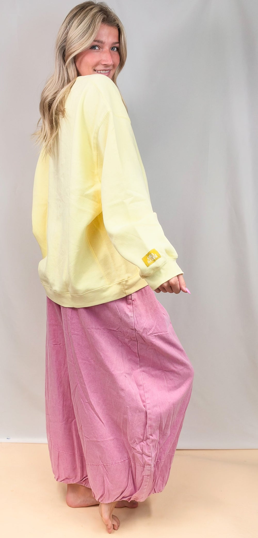 Woman shown from side view wearing pale yellow sweatshirt and pink balloon pants on plain background