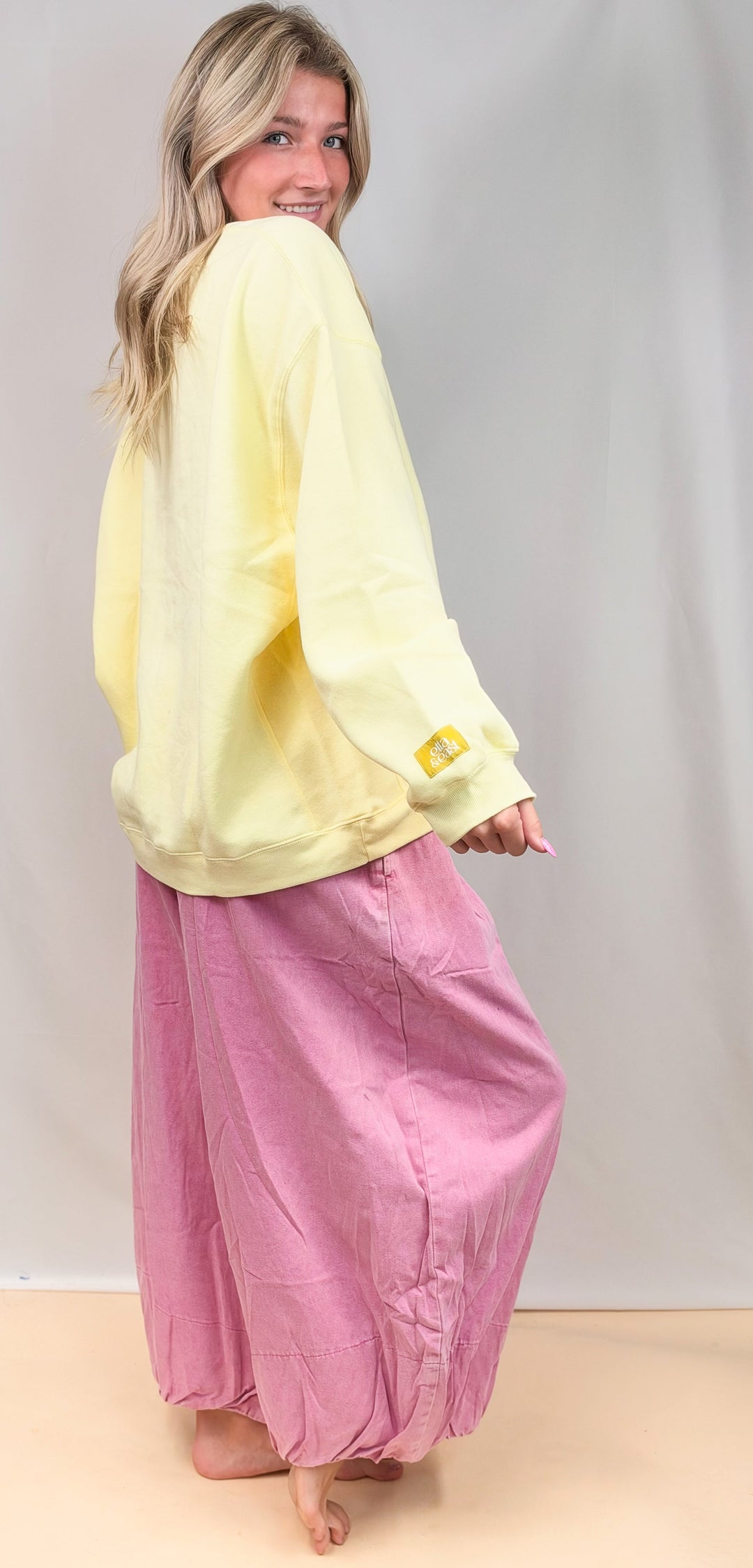 Woman shown from side view wearing pale yellow sweatshirt and pink balloon pants on plain background