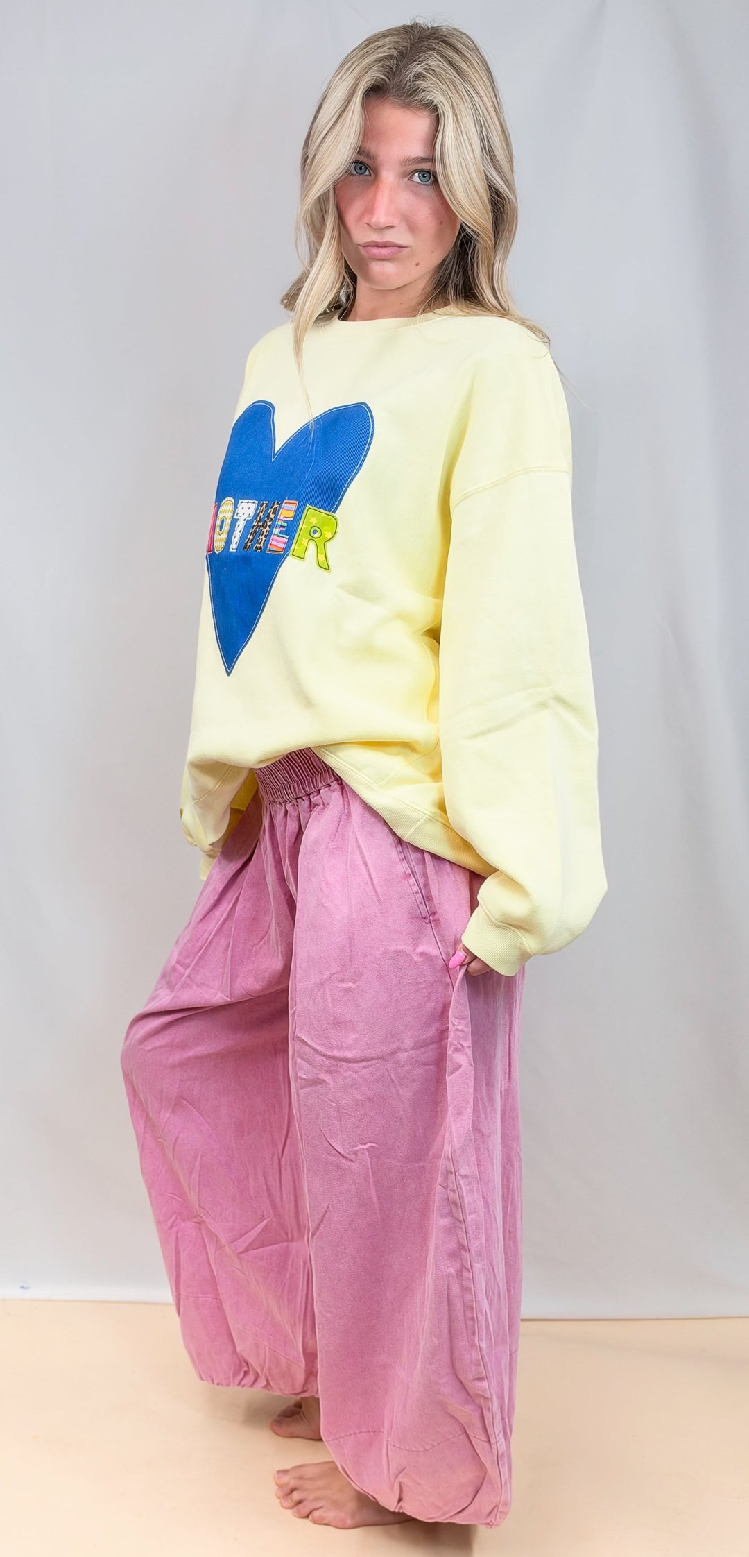 A woman stands barefoot against a light gray studio backdrop, wearing an oversized pale yellow sweatshirt with a large blue heart graphic and the word &