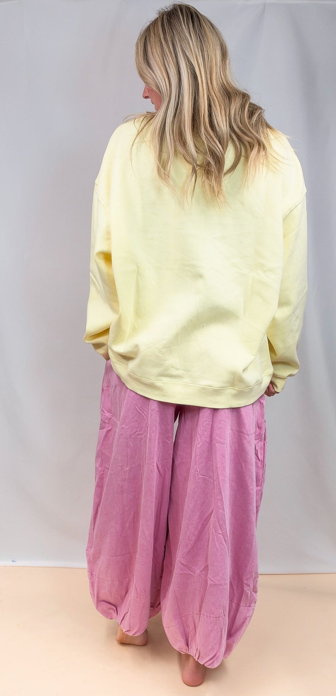 A woman shown from behind wearing a pale yellow oversized sweatshirt and loose pink harem-style pants, standing against a plain light gray studio backdrop.