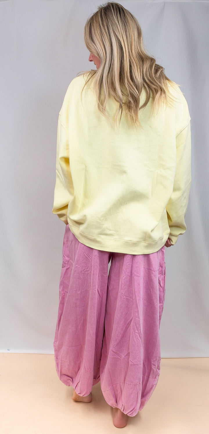 A woman shown from behind wearing a pale yellow oversized sweatshirt and loose pink harem-style pants, standing against a plain light gray studio backdrop.