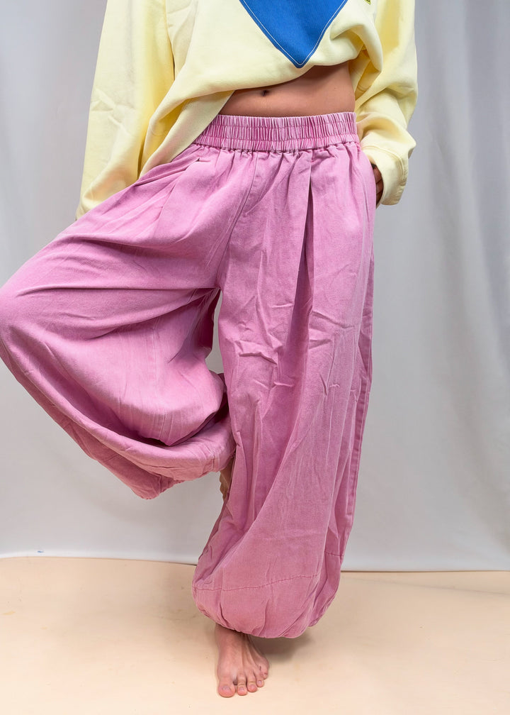 A person wearing loose, baggy pink harem-style pants with an elastic waistband, paired with a pale yellow and blue sweatshirt, standing barefoot against a light gray backdrop.
