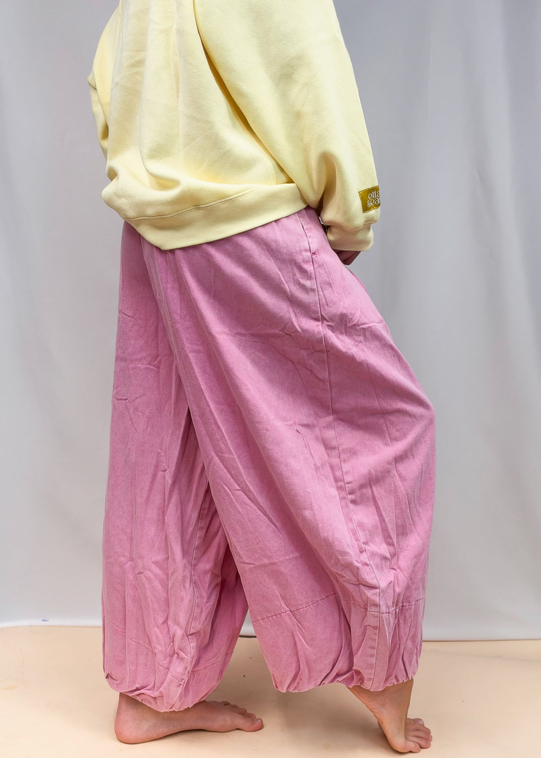 A person wearing loose, baggy pink harem-style pants and a pale yellow sweatshirt, standing barefoot against a plain light backdrop.