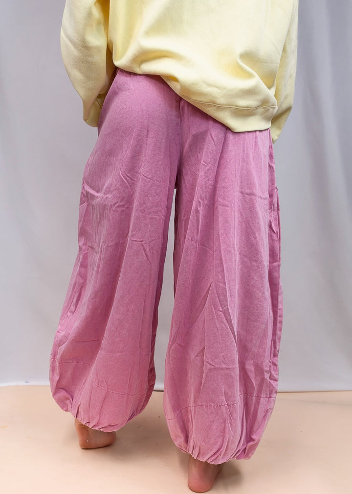 A person shown from the back wearing loose, wide-leg pink pants with a pale yellow top, standing barefoot against a plain light backdrop.