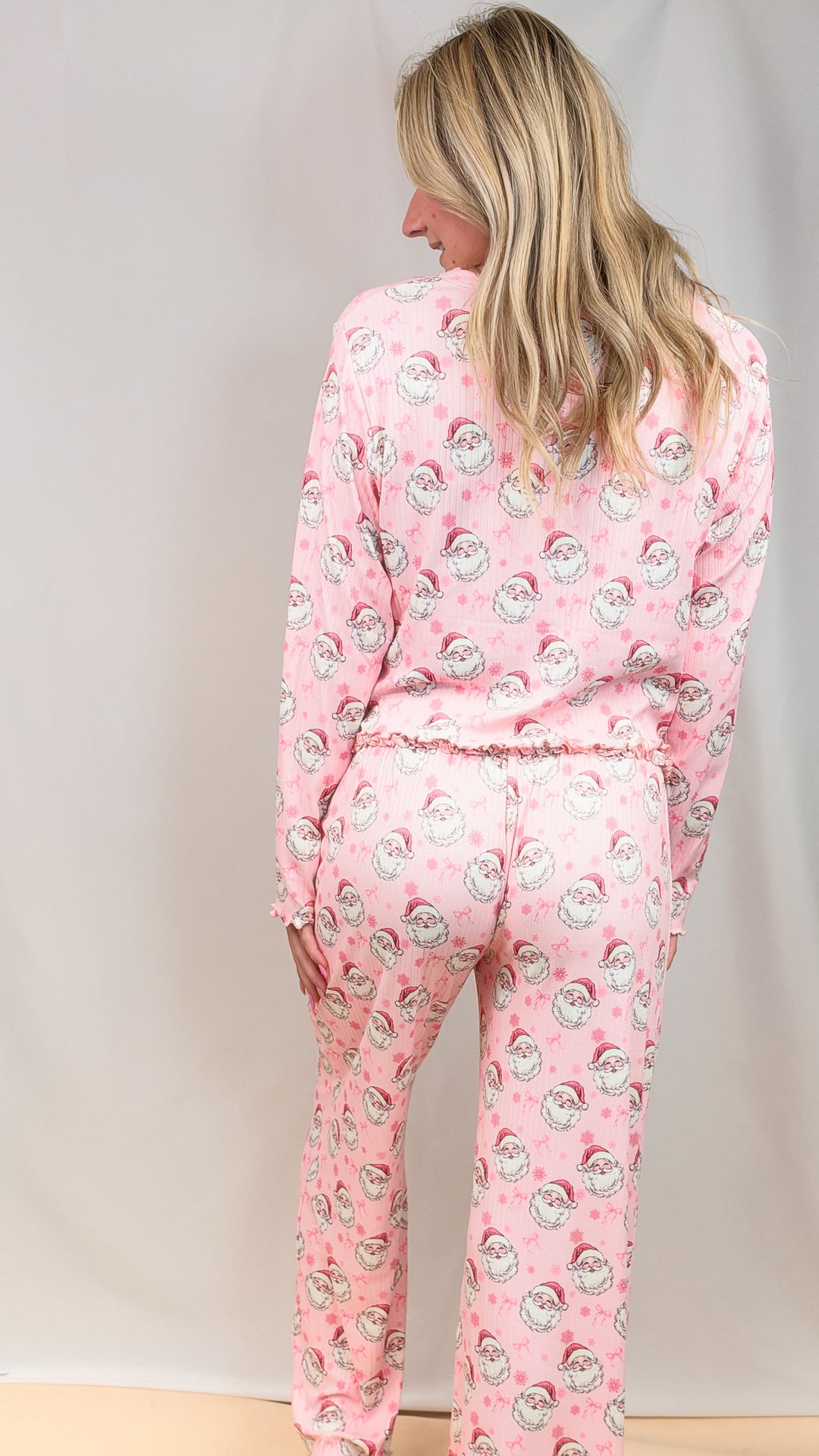Woman shown from back wearing pink christmas pajamas with vintage santa faces and bows on plain background