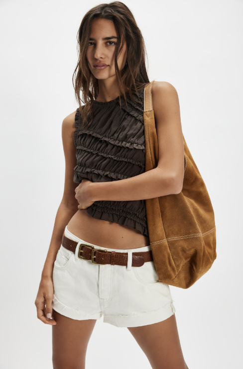 Woman wearing a brown sleeveless top and white shorts, holding a tan bag on a white background