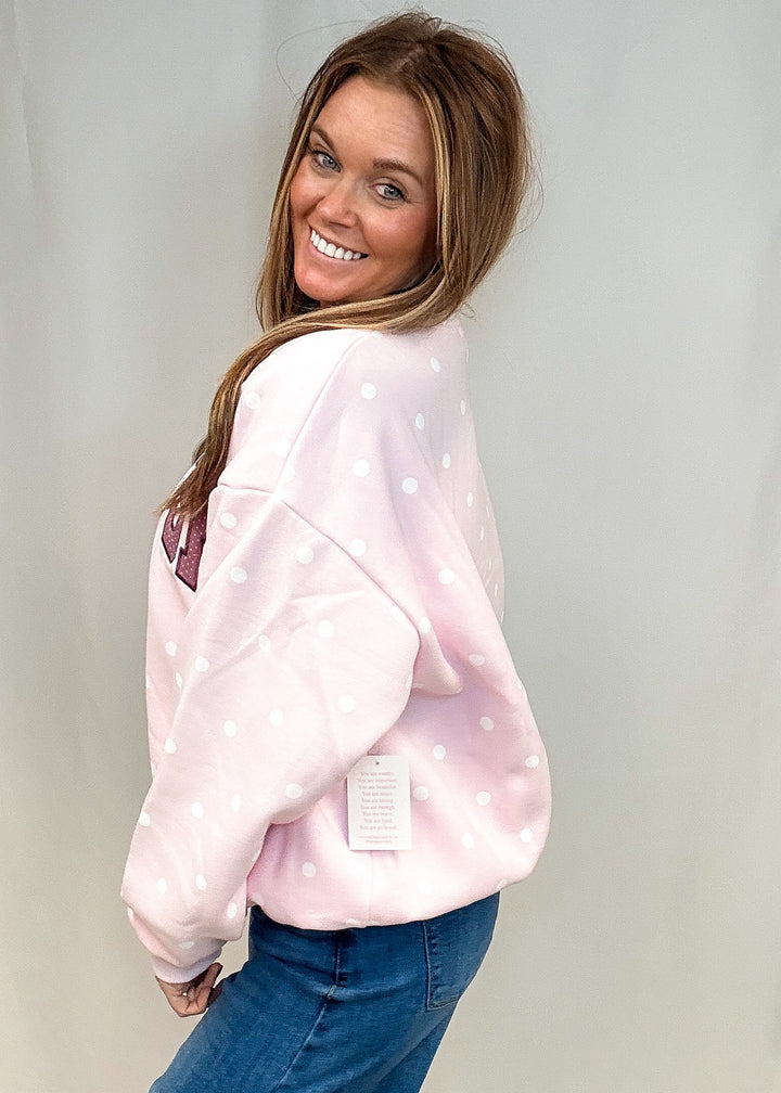 A woman wearing a light pink sweatshirt with white polka dots and blue jeans, posing in front of a white studio backdrop.
