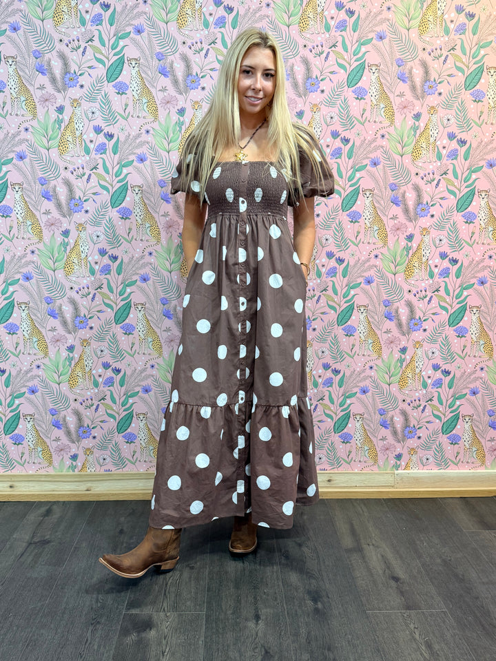 Woman wearing a brown polka dot dress standing in front of a patterned wall.