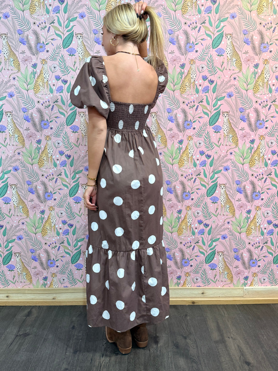 Woman wearing a brown dress with white polka dots against a patterned wall.