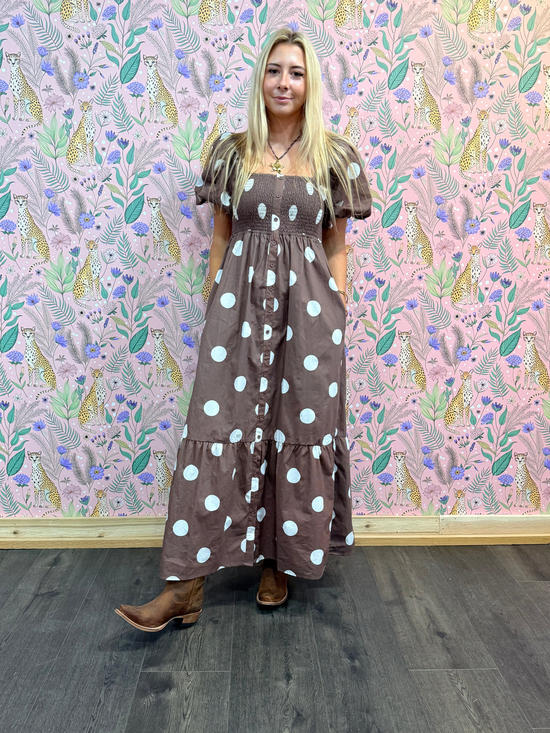 Woman wearing a brown dress with white polka dots standing in front of a patterned wall.