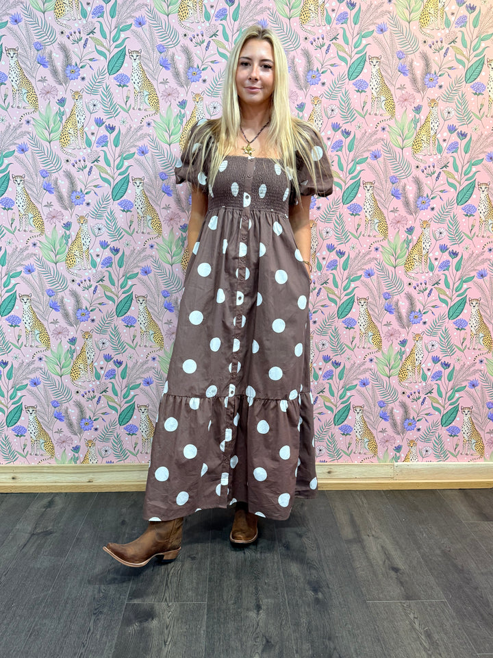 Woman wearing a brown dress with white polka dots standing in front of a patterned wall.
