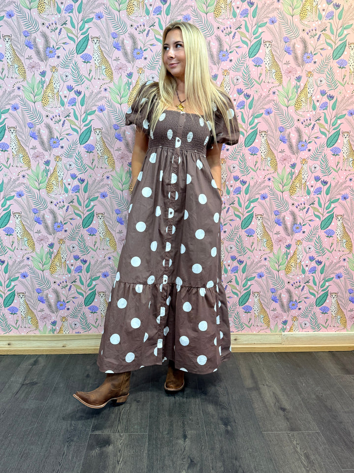 Woman wearing a brown dress with white polka dots against a patterned wall.