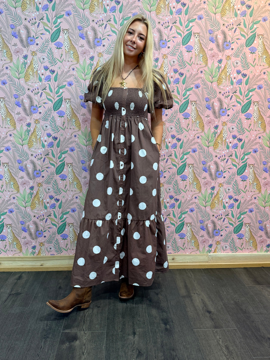 Woman in a brown polka dot dress standing against a patterned wall.