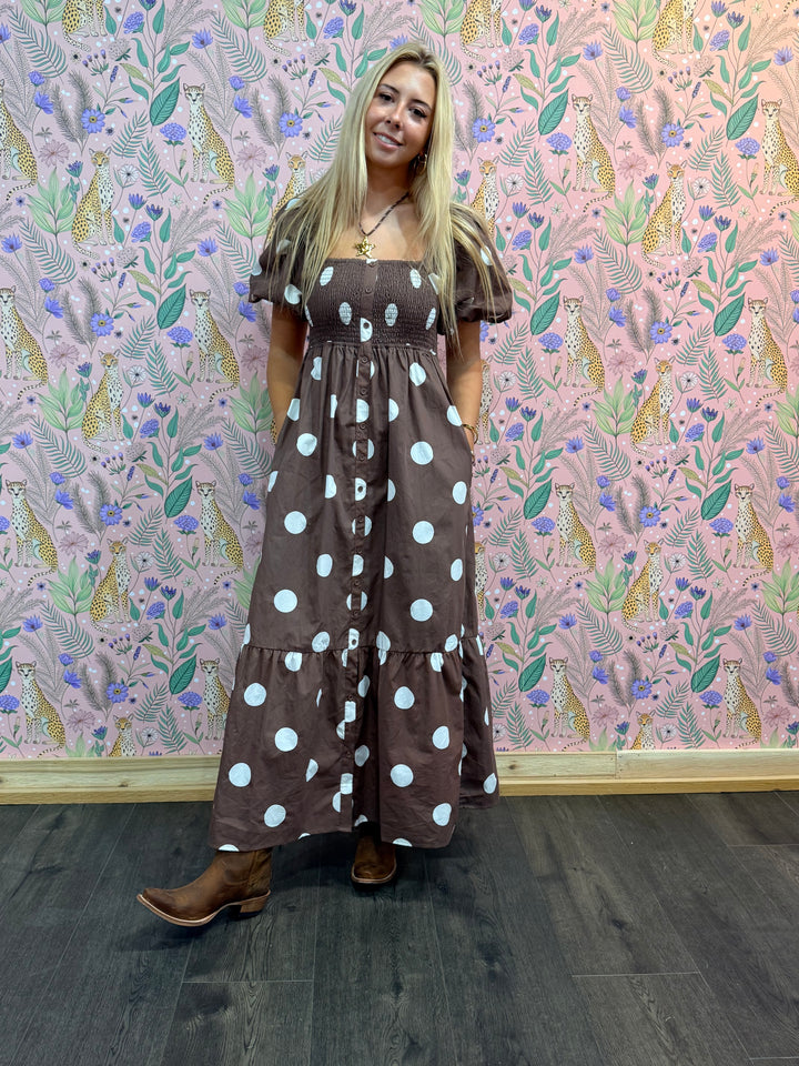 Woman in a brown polka dot dress standing against a patterned wall.