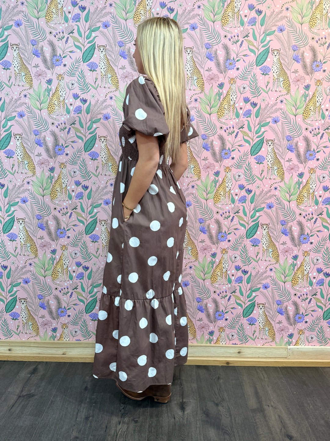 Woman in a brown dress with white polka dots standing against a pink floral wallpapered wall.
