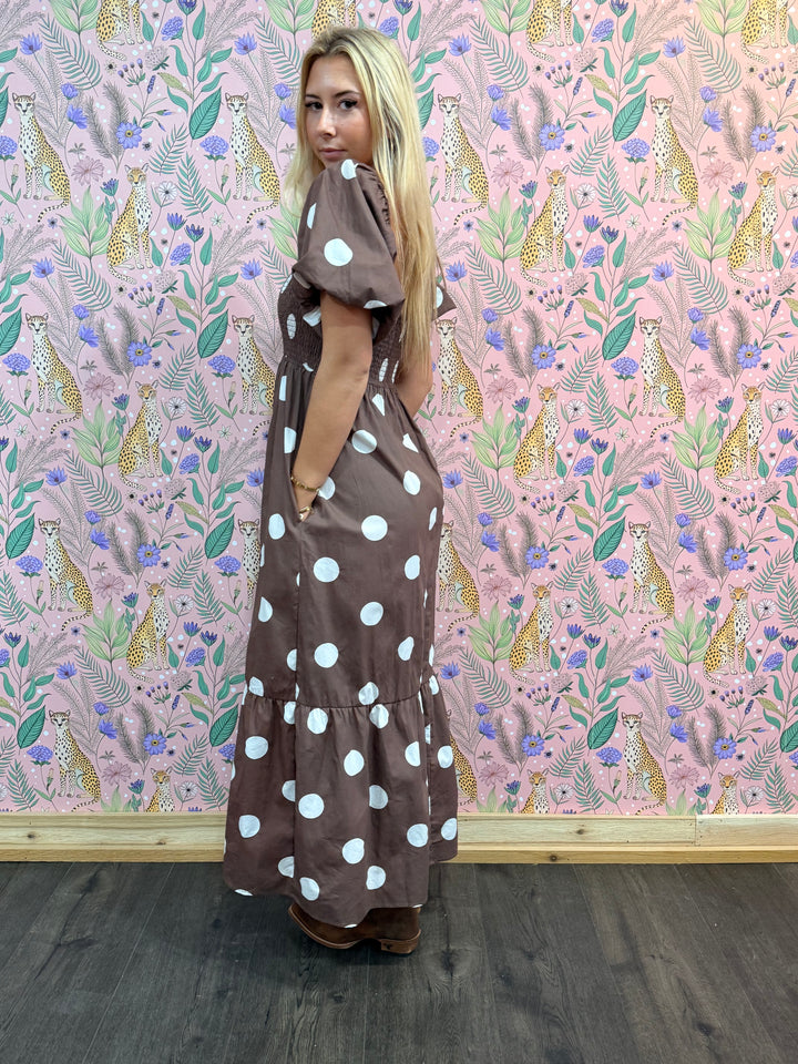 Woman wearing a brown dress with white polka dots against a pink floral patterned wall.