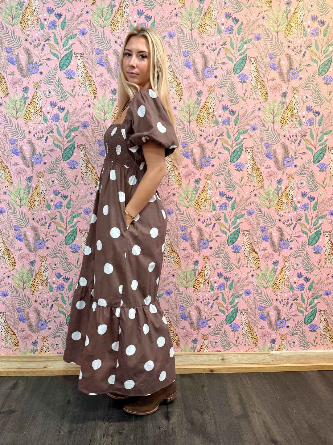 Woman in a brown dress with white polka dots standing against a pink floral and animal patterned wall.