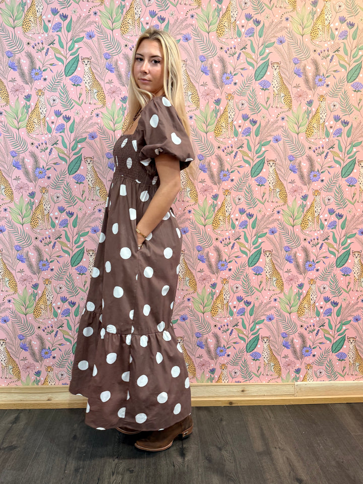 Woman in a brown dress with white polka dots standing against a pink floral and animal patterned wall.