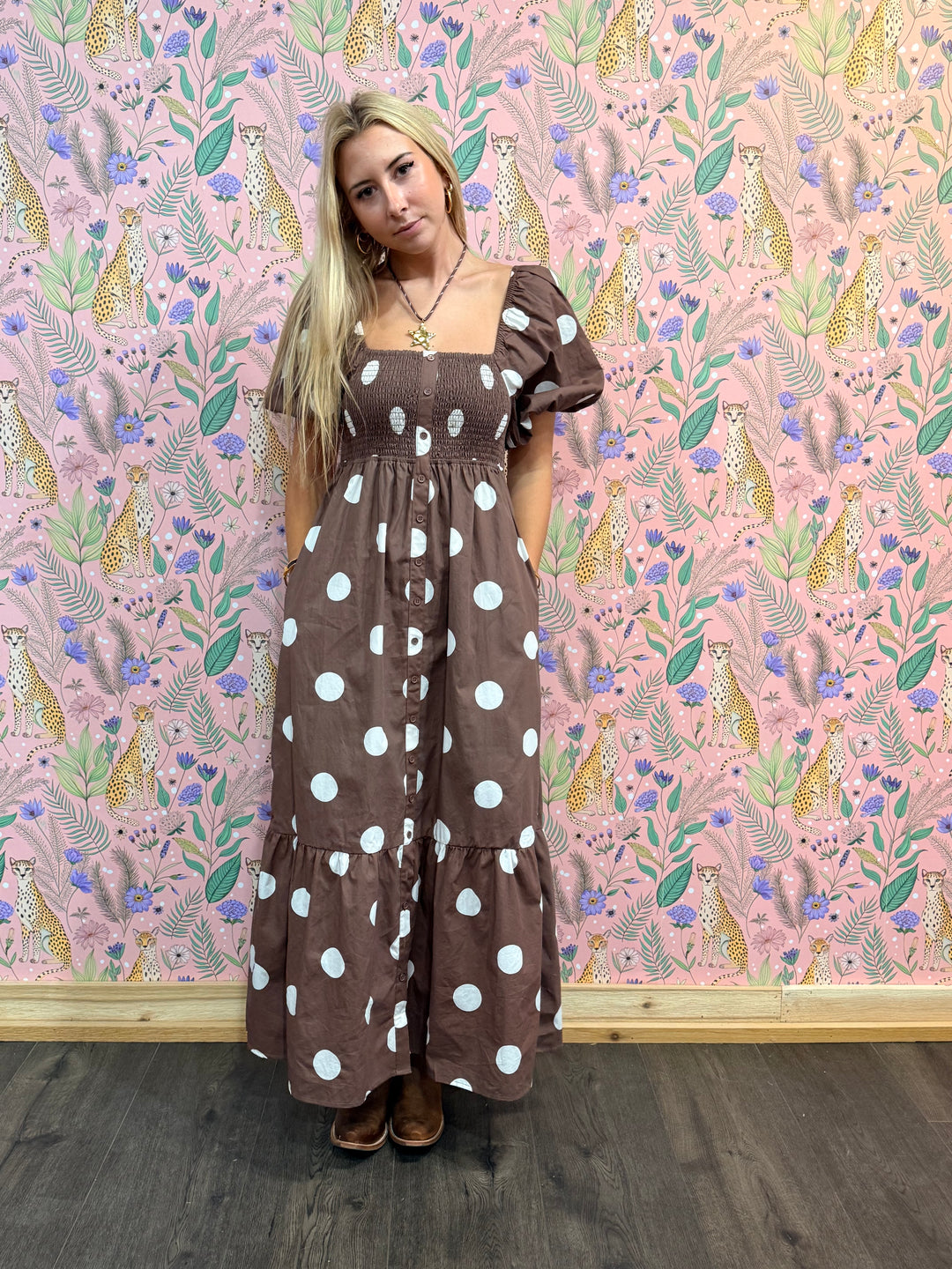 Woman wearing a brown dress with white polka dots against a pink wallpapered wall.