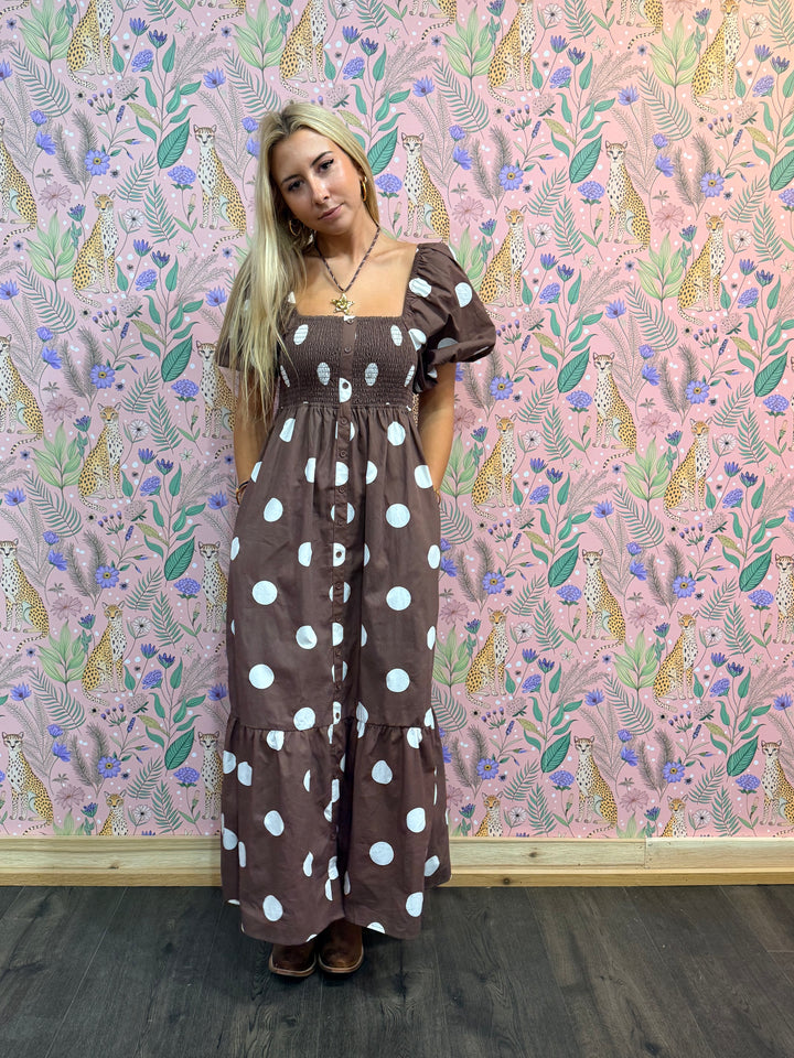 Woman wearing a brown dress with white polka dots against a patterned wall.
