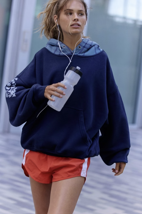 A woman wearing an oversized navy blue hoodie with a plaid hood and white sleeve embroidery, paired with red athletic shorts, walking outdoors while holding a water bottle and wearing white earphones.