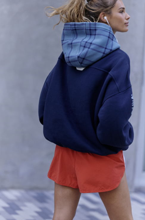 A woman wearing an oversized navy hoodie with a light blue plaid hood lining, paired with rust-orange athletic shorts, photographed outdoors against a gray wall.