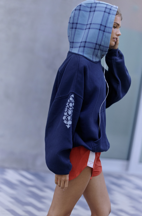 A woman wearing a navy blue hoodie with a light blue plaid hood, walking outdoors. The hoodie features a white floral graphic on the sleeve.