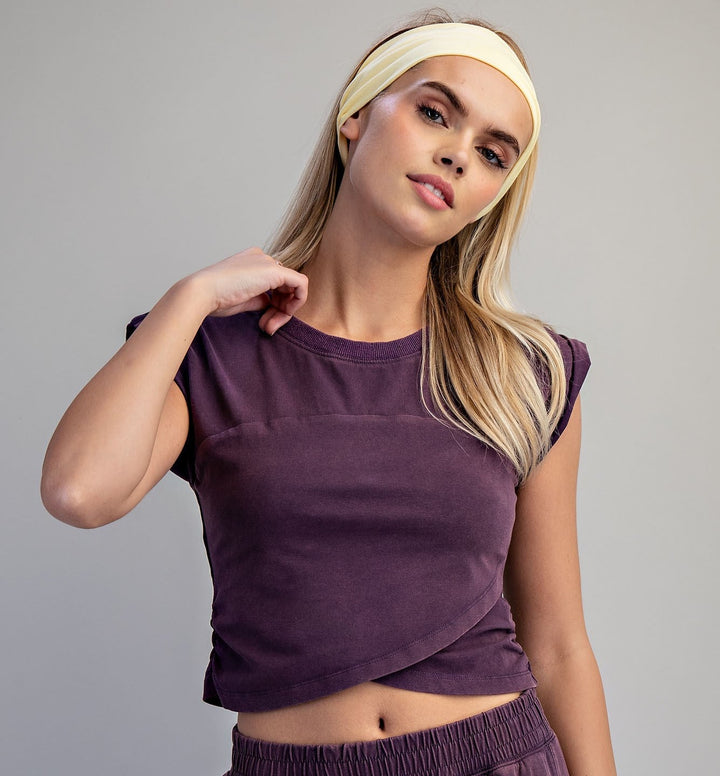 A woman modeling a matching two-piece lounge set in deep purple, featuring a short-sleeve crop top and matching skort, paired with a cream-colored headband against a solid gray studio background.