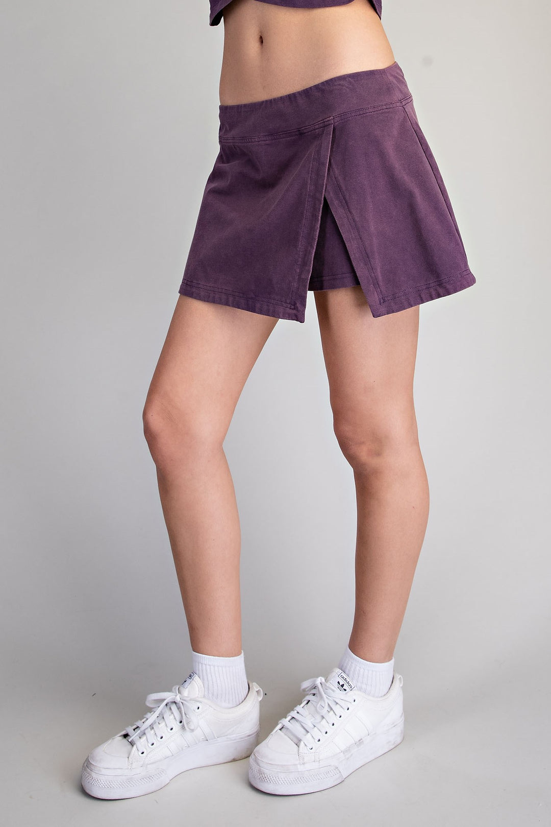 A person wearing a purple wrap-style mini skirt with side slits, paired with white crew socks and white platform sneakers, photographed against a light gray studio background.