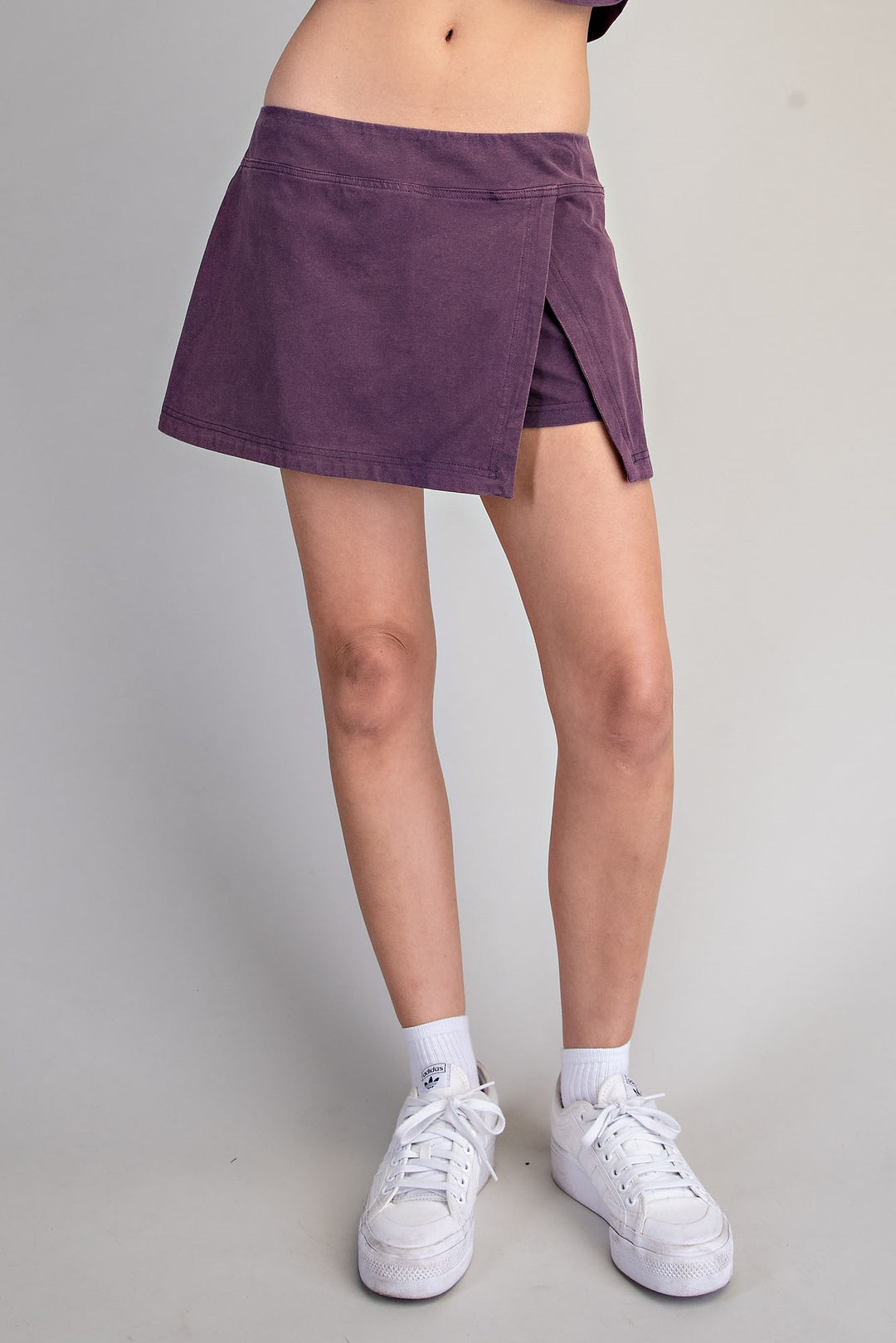 A person wearing a short wrap-style skirt in a muted purple corduroy-like fabric, paired with white socks and white sneakers, photographed against a plain light gray studio background.