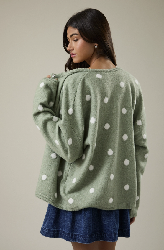 A woman wearing a sage green knit sweater with white polka dots, shown from a side-back angle against a plain beige studio background.