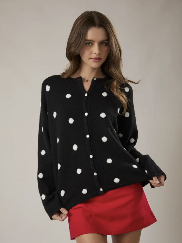 A woman modeling a black knit cardigan sweater with white polka dots and a red mini skirt, photographed against a plain light beige studio background.