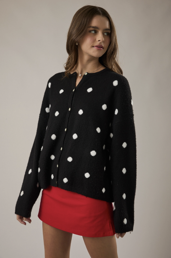 A woman modeling a black fuzzy cardigan with white polka dots and a red mini skirt against a plain beige studio background.
