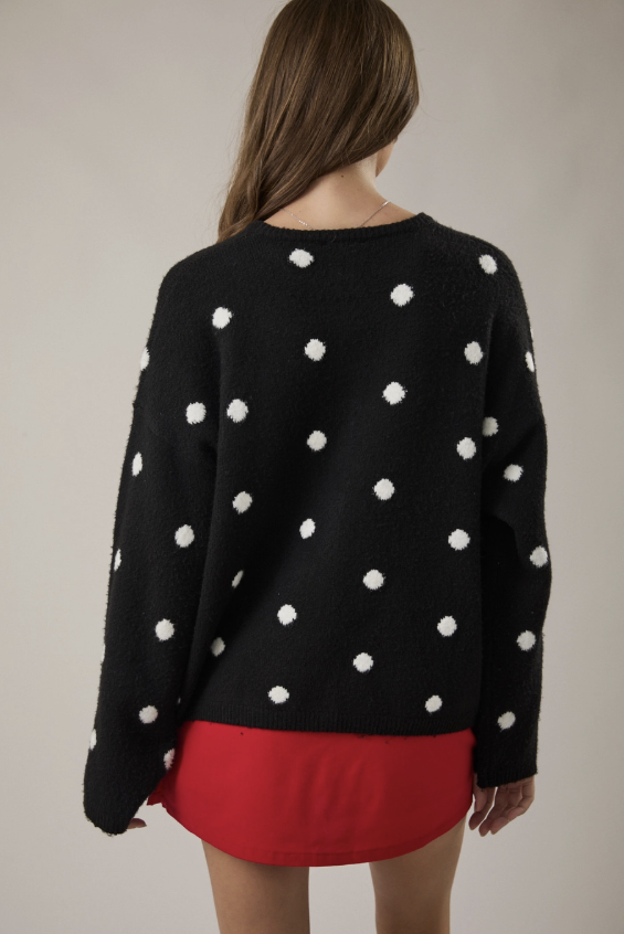 A woman shown from the back wearing a black fuzzy knit sweater with white polka dots and a bright red skirt, against a plain light gray studio background.