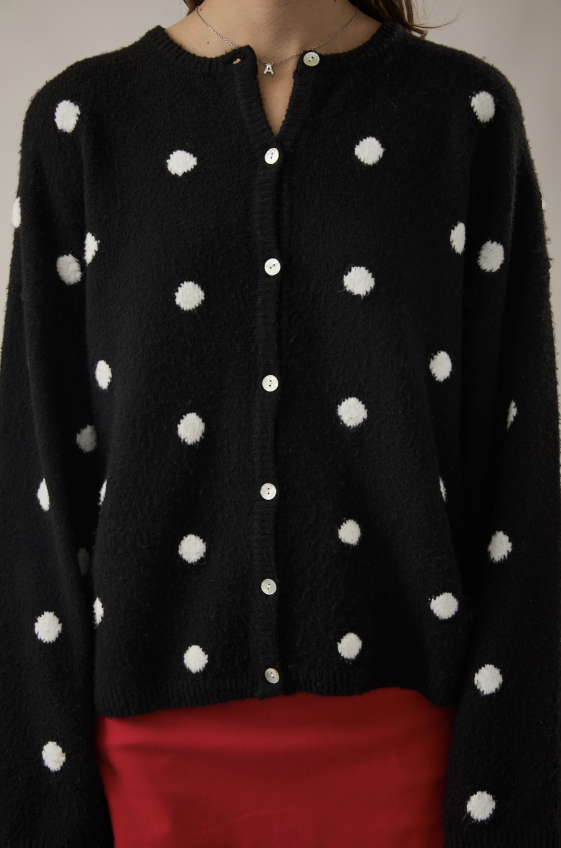 A close-up of a person wearing a black fuzzy knit cardigan with white polka dots and white buttons, paired with a red skirt or bottom.