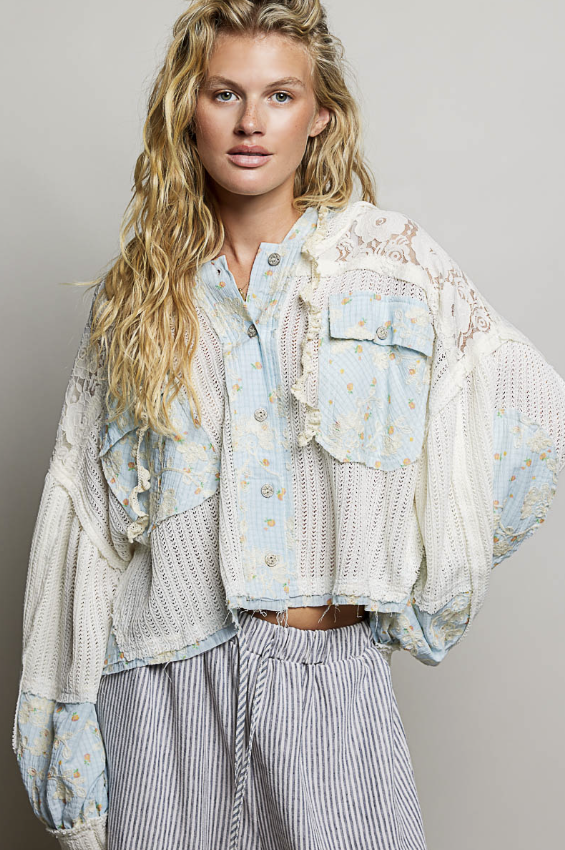 A woman modeling a bohemian-style cropped button-up shirt jacket made of light blue floral lace and cream crochet mesh, paired with striped trousers, against a neutral gray background.