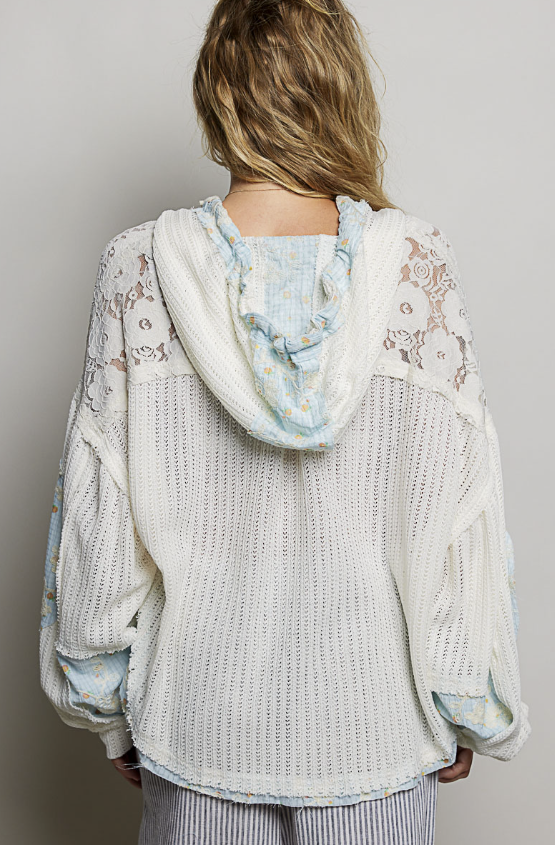 A woman viewed from behind wearing a cream-colored waffle-knit hooded sweater with light blue and white lace shoulder panels and a ruffled light blue hood.