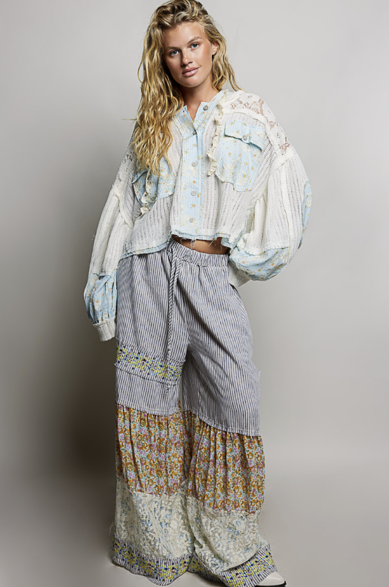 A woman modeling a bohemian-style outfit featuring a cream and light blue striped cropped jacket with ruffles, matching striped and floral wide-leg pants, and white sneakers, set against a plain light gray studio background.