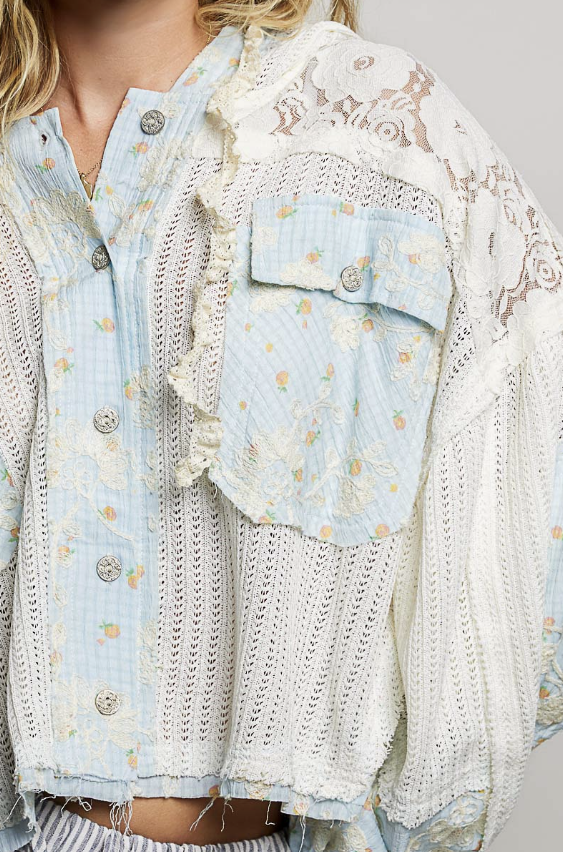 A close-up of a bohemian-style light blue and white floral button-up shirt jacket, featuring a lace overlay, ruffled trim, and a chest pocket with a decorative button.