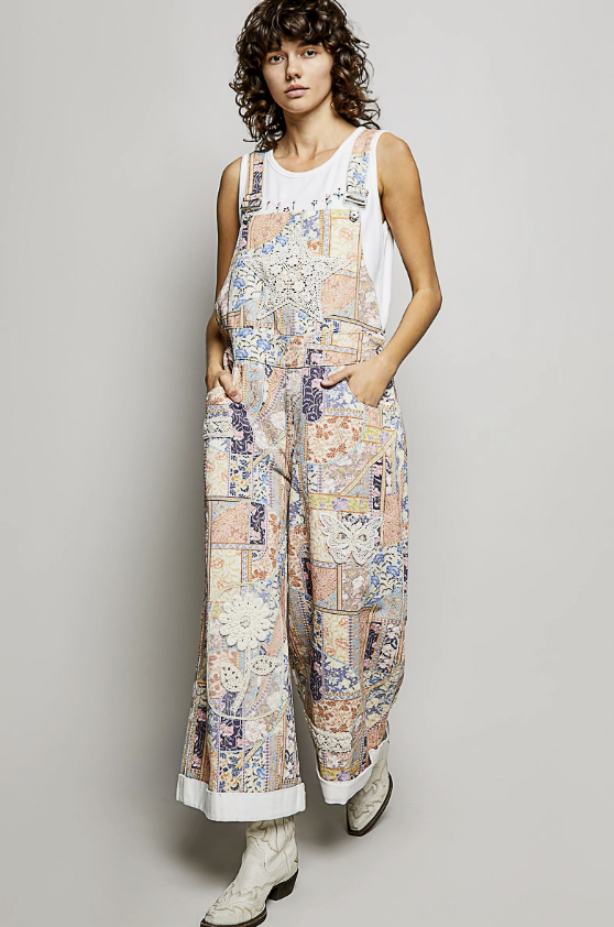 A model stands against a light gray studio background wearing a sleeveless white tank top and matching wide-leg overall jumpsuit with a multicolor patchwork print in beige, blue, pink, and cream. The jumpsuit has a bib front, pockets, and cuffed hems, paired with white lace-up boots.