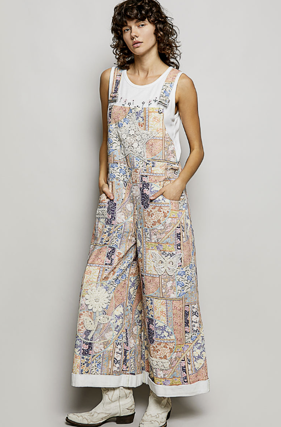 A model wearing a pair of wide-leg canvas overalls with a vintage patchwork print in beige, blue, and orange, styled over a white tank top and white boots against a light gray studio background.