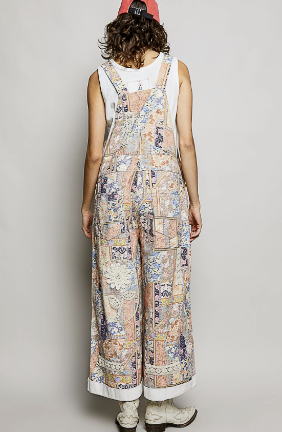 A model shown from the back wearing a white sleeveless top and light blue patchwork wide-leg overalls, paired with a red cap and white boots, against a neutral gray background.
