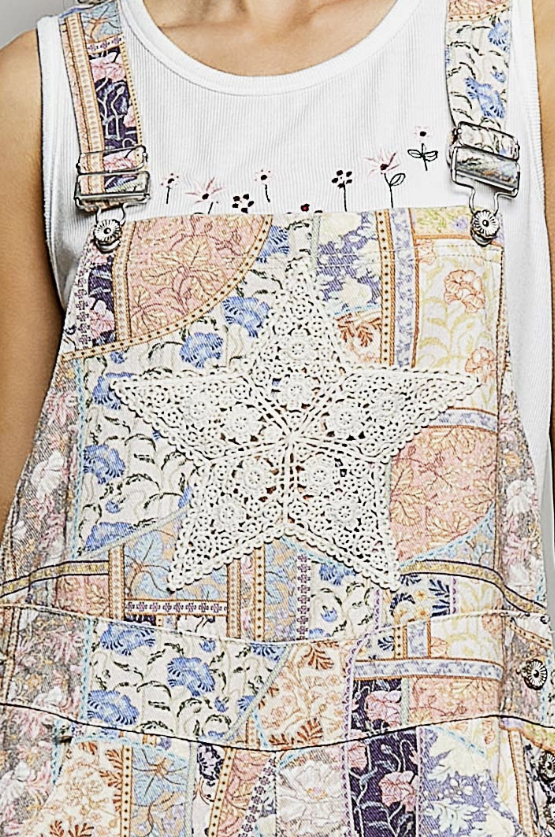 A close-up of a person wearing patchwork-style patchwork overalls with floral and lace-like printed panels, a large white star motif at the center, and adjustable metal-clip straps over a white sleeveless top.