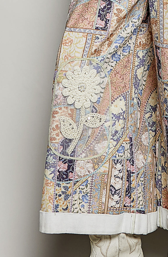 Close-up of a patchwork-patterned jacket or coat with floral embroidery, a large embroidered flower motif, and a white ruffled hem, shown against a plain light gray background.