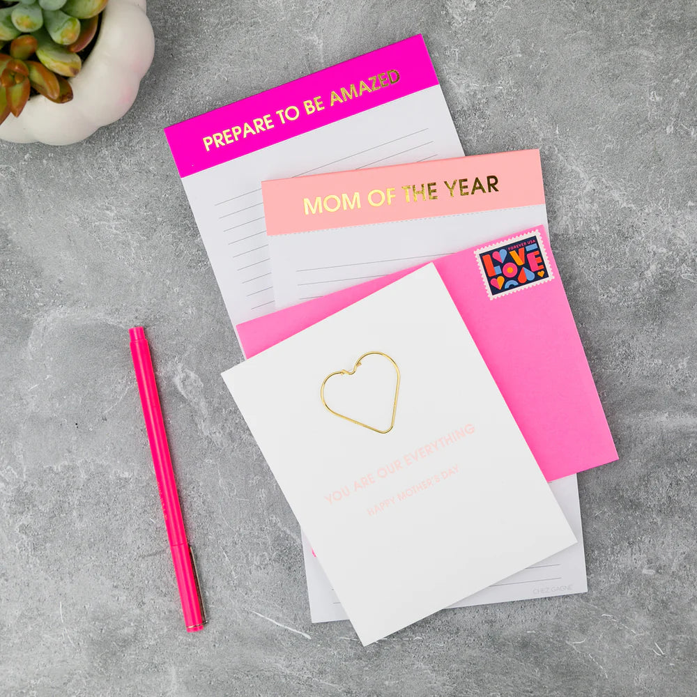 Notebook with 'Mom of the Year' text, pink pen, and heart-shaped paper clip on a gray surface.